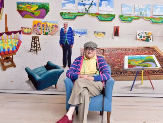 David Hockney seduto su poltrona azzurra nel suo studio d arte circondato da grandi tele prospettiche e paesaggi colorati