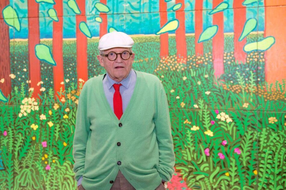 David Hockney in cardigan verde e cravatta rossa davanti al dettaglio di un suo grande dipinto naturalista boschivo con alberi rossi