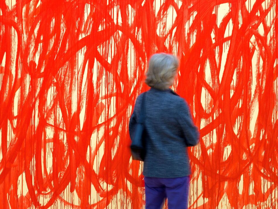 Donna di spalle con giacca grigia osserva un immenso quadro astratto coperto da un groviglio di linee rosse colate