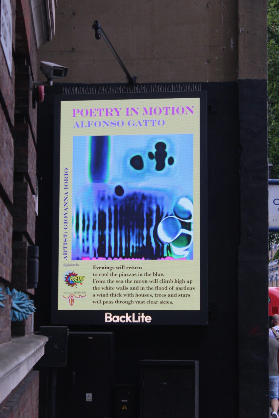 Cartellone luminoso verticale "BackLite" installato sulla facciata esterna di un edificio in mattoni a Londra. Lo schermo mostra il titolo "Poetry in Motion - Alfonso Gatto" e uno spettrogramma vocale azzurro e blu con forme astratte scure, seguito dai versi della poesia "Evenings will return" e dal logo della Fondazione Alfonso Gatto.