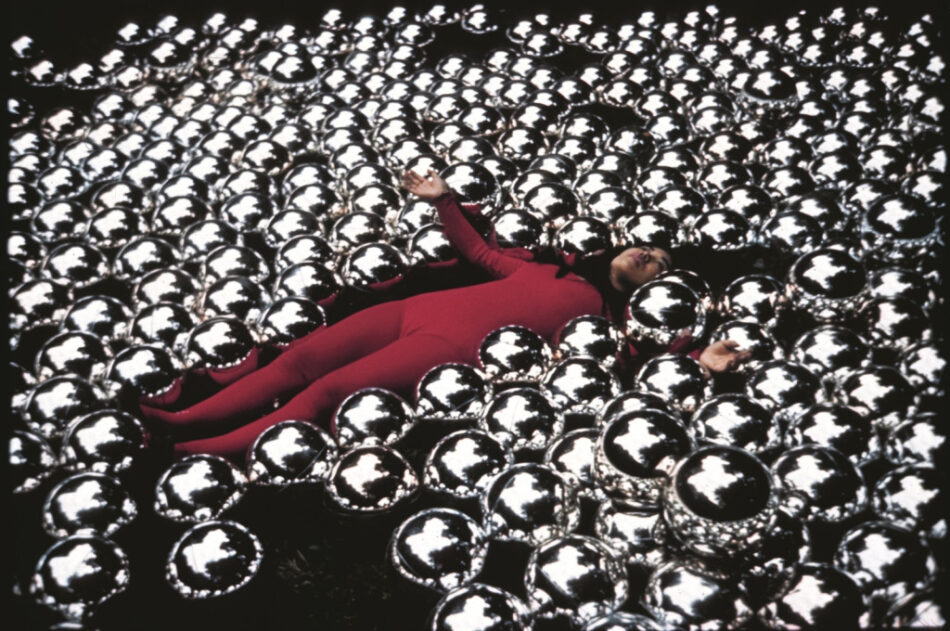 Yayoi Kusama in tuta rossa distesa su un prato verde coperto da centinaia di sfere metalliche riflettenti.